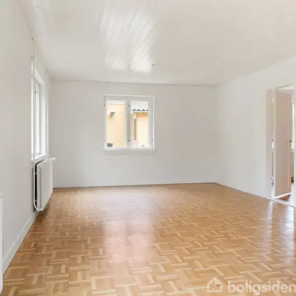 Et lyst rum med parketgulv og hvide vægge. To vinduer giver naturligt lys, og der er døre til tilstødende værelser. Radiator langs væggen til venstre og intet møblement.