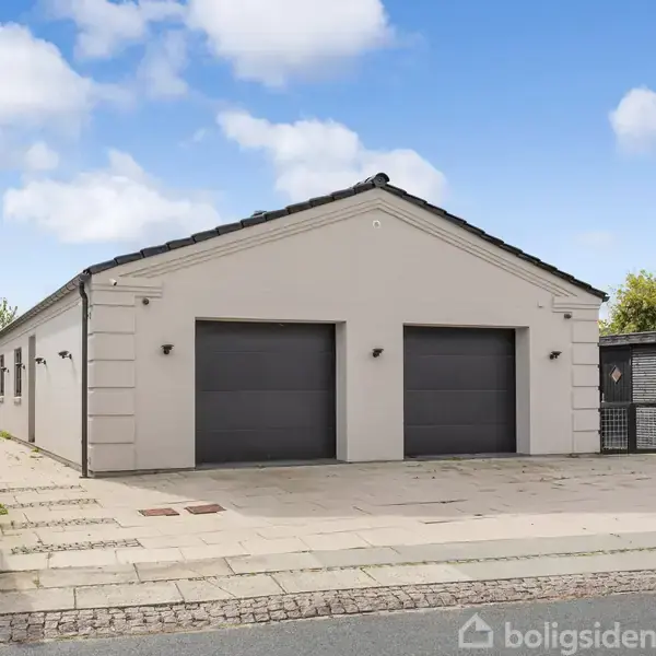 En bygning med to store garageporte, står stille på en flisebelagt indkørsel. Ved siden er en sort skur. Konteksten er et boligområde med klare himmel og grønne buske. Skilt: "Ny bolig! Se alle boliger på nybolig.dk".
