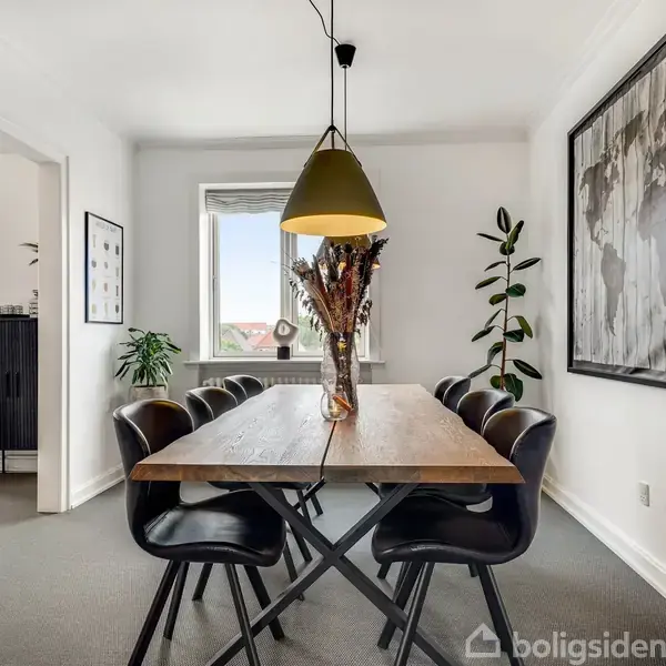 Et træbord med seks sorte stole står i et lyst spisestue. En vase med tørrede blomster pryder bordet. Over bordet hænger en lampe, mens et stort billede hænger på væggen.