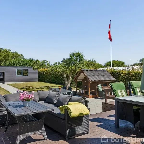 Terrassemøbler står arrangeret på en træveranda med en parasol, omgivet af en græsplæne og en lille legeplads. En dansk flagstang står i baggrunden under en blå himmel.