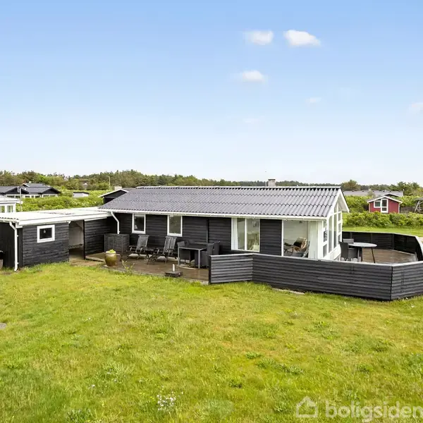 Et sortmalet sommerhus står i en grøn have med en terrasse foran, hvor der er placeret havemøbler. I baggrunden ses flere huse og en skov under en lys himmel.