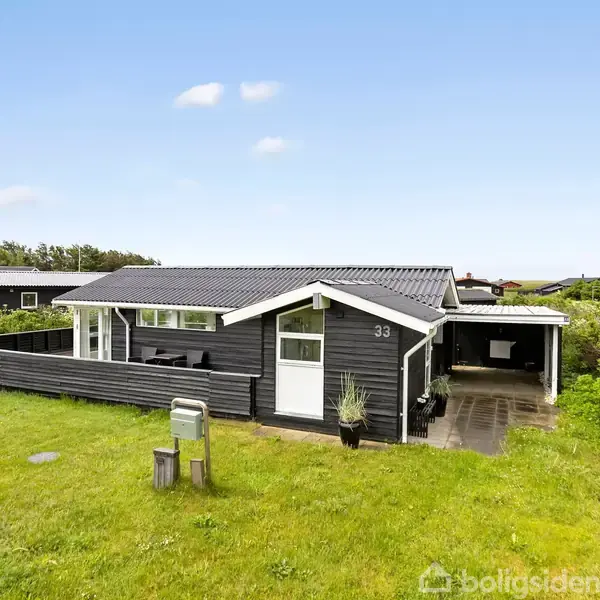 Stue med moderne indretning; en sofa og stol foran store vinduer. Til højre, et hjørne med en reol, lampe og dekorationer på en hvidmalet væg.