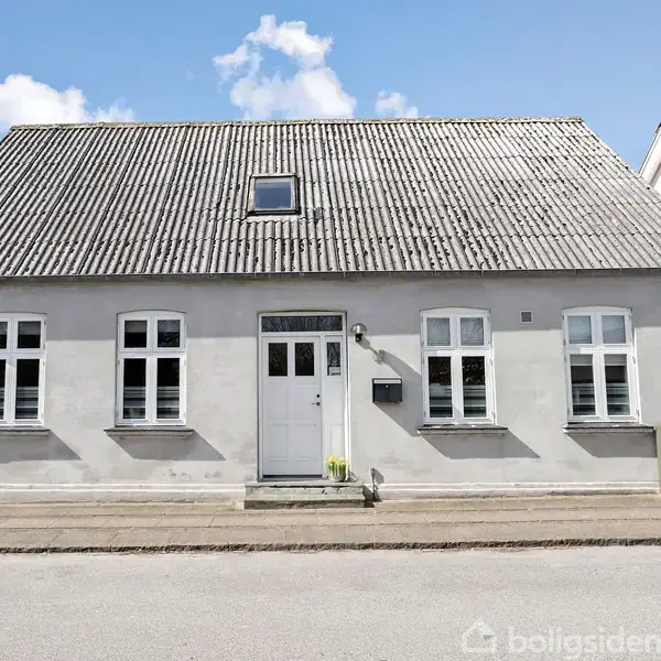 Et hvidt, traditionelt hus står stille på en rolig gade, omgivet af andre bygninger. Det har flere vinduer med sprosser og en mørkgrå tag. Himlen er let skyet.