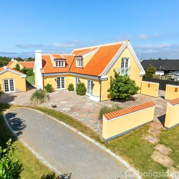 Gul villa med rødt tag står omgivet af en indhegnet have og belægningssten. Omkringliggende bebyggelse ses i baggrunden under en klar blå himmel.