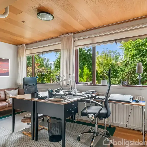 Kontor med skrivebord fyldt med papirer, computere og en sort kontorstol. Bagved står en brun lædersofa. Store vinduer viser en have med frodig beplantning. Væggene er lyse, og en bogreol fyldt med mapper ses til højre.