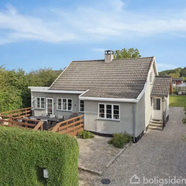 En gråmalet villa med mørkt tag står ved siden af en grusvej, omgivet af hække og træer. Foran huset er en terrasse med møbler, omkranset af et trægelænder.