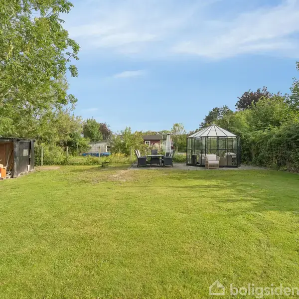 Et drivhus står i en have omgivet af træer og buske. Til venstre ses et skur under et stort træ. Der er blå himmel og sollys.