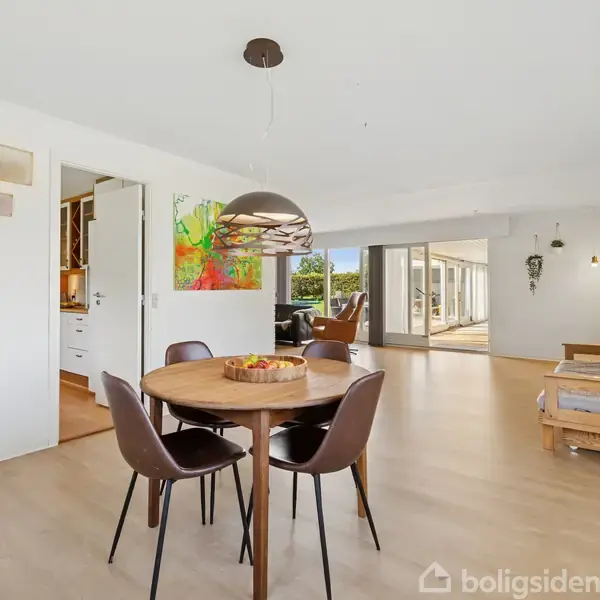 Rundt spisebord med fire stole står i et lyst rum med trægulv. En farverig vægdekoration hænger på væggen, og glasfacader åbner op mod en terrasse udenfor.
