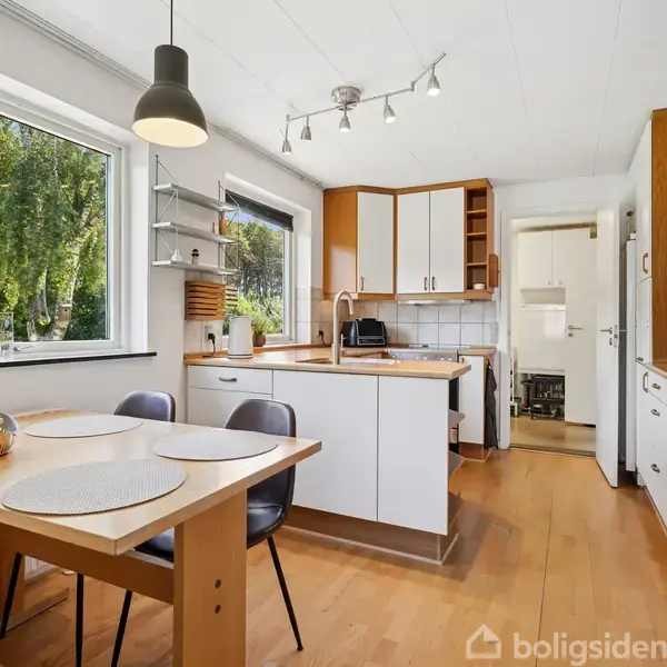 Et køkken med træbord og fire stole ved vinduet, køkkenø med vask, hvide skabe og apparater, samt store grønne træer udenfor i dagslys.