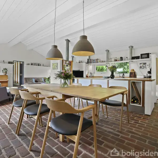 Spisebord med seks stole står i et rum med murstensgulv. Store lamper hænger over bordet, mens en grå sofa og hylder udsmykker baggrunden. Lyst interiør dominerer.