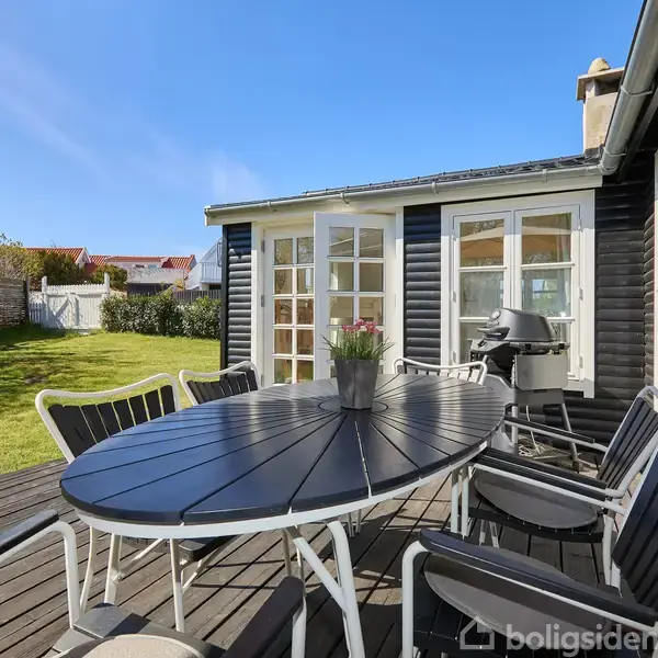 Et ovalt bord med stole står på en træterrasse foran et sortmalet hus med mange vinduer. I baggrunden ses en velplejet have og en høj mur.