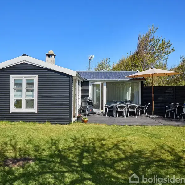 Et sort træhus med hvide vinduer står på en grøn græsplæne. Foran huset er der en terrasse med havemøbler og en parasol. Solrigt vejr og omgivet af buske og træer.