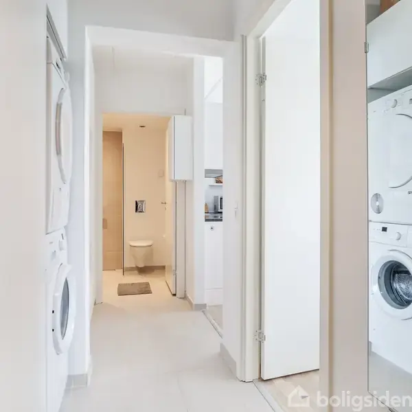 Vaskemaskiner og tørretumblere er stablet i et smalt vaskerum. I baggrunden ses en dør til et badeværelse med toilet og lyse fliser.