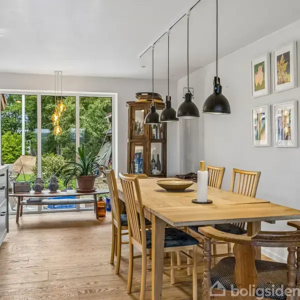 Et træbord med fire stole står i et moderne køkken med hvide vægge. Bordet har en lysestage og skåle. Store vinduer i baggrunden giver udsigt til en grøn have.