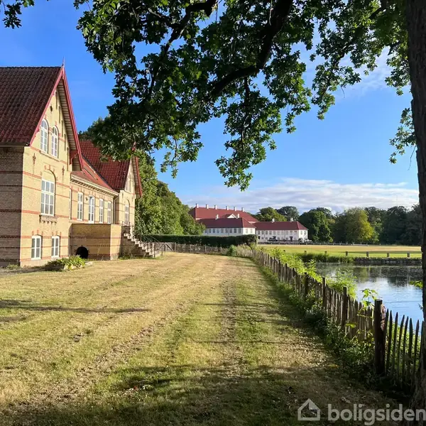 En stor bygning med rødt tegltag står ved siden af en græsplæne og en sø. En træramme med grene omkranser området, og der er åbent, klart vejr.