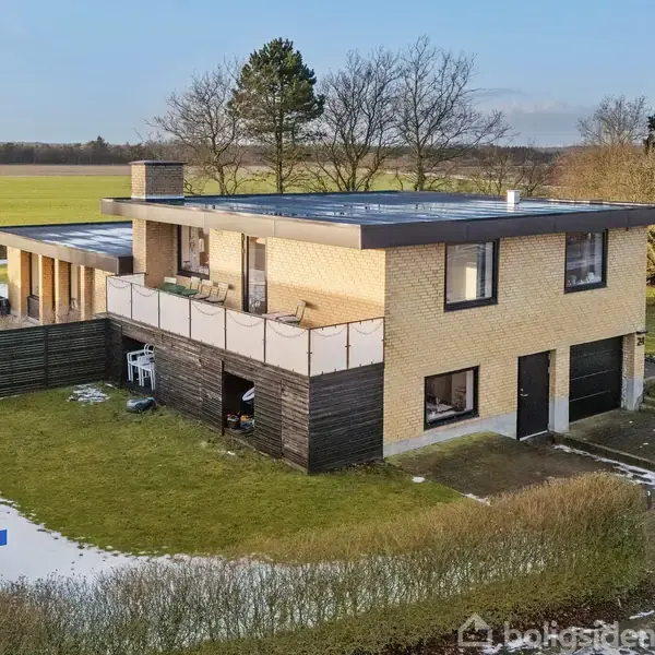 To-etagers hus med fladt tag og gul murstensfacade, står på grøn græsplæne. Snepletter dækker jorden, omgivet af marker og træer. Vejskilte: "Rønnealle" og "E2-8".