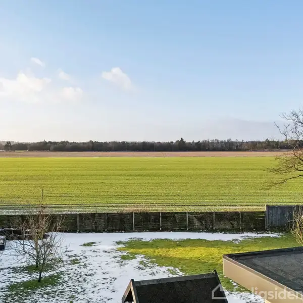 Grønt markstræk strækker sig mod horisonten, omgivet af træer. Forgrundens have viser en blanding af græs og smeltende sne, mens bygninger sidder ved siden af. Himmelen er lys.