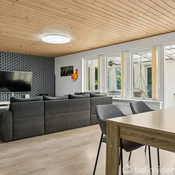 En stor, grå sofa står i en stue foran et vægmonteret fjernsyn; lyse træpaneler pryder loftet, mens vinduer i baggrunden lukker sollys ind og afslører en overdækket terrasse.