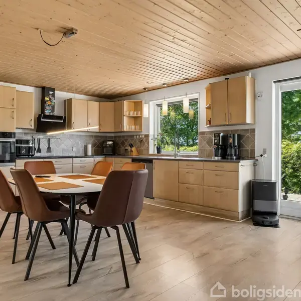 Køkken med træloft har moderne bord og seks stole. Køkkenet har skabe, ovn og køleskab. Store vinduer og døre giver udsigt til grøn have. Skinnende træbelægning fuldender rummet.