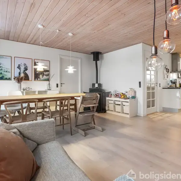 En spisebordsscene med stole står i et rummeligt rum med trægulve og træloft. Hængelamper lyser op, og en brændeovn er placeret i hjørnet. Væghængte billeder ses bagved.