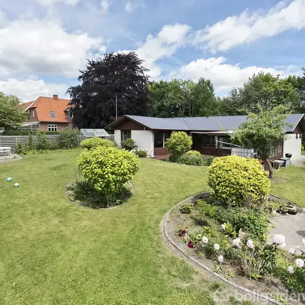 En velplejet have med blomsterbede og buske omgiver et enkelt hvidt hus med sort tag. Grønne træer og en blå himmel danner baggrunden.
