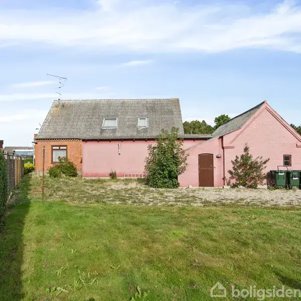 Et lyserødt hus med skråtag står på en velholdt græsplæne; omgivet af buske og træer i et beboelseskvarter. Flere affaldscontainere ses ved siden af huset.