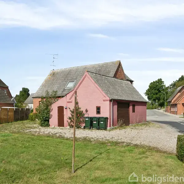 Et lyserødt hus står stille i et grønt kvarter med andre huse omkring. Foran huset er der tre affaldscontainere, og en vej løber langs siden af ejendommen.