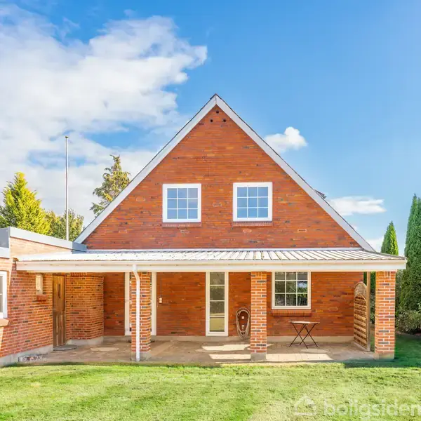 Rødstenshus med to etager og to vinduer på førstesalen. Huset har en overdækket terrasse og står i en velplejet have omkranset af buske og træer under en blå himmel.