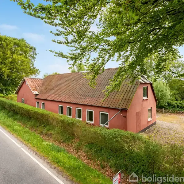 Rødt hus med tag af tegl står langs en landevej. Omkranset af grønne træer og buske under en blå himmel.