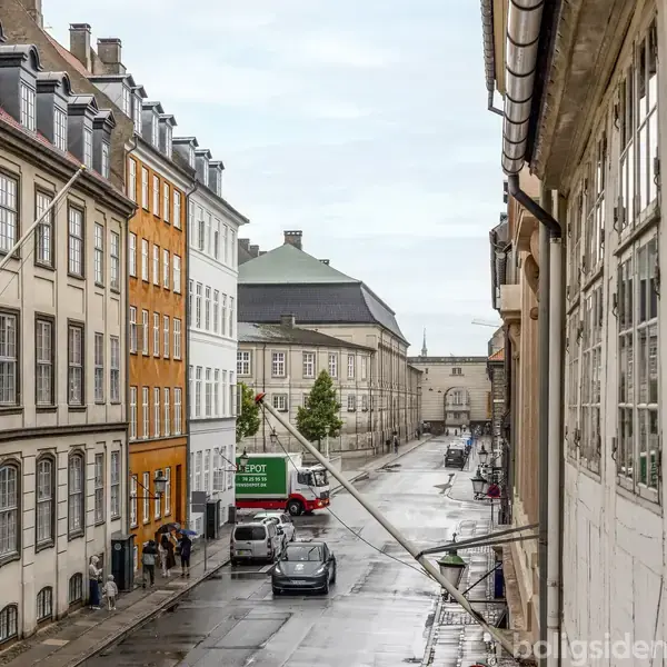 Flere biler kører på en smal, brostensbelagt gade mellem rækker af gamle bygninger i forskellige farver, herunder en markant orange. Skiltet på en lastbil siger: "Depot." Vejret er overskyet.