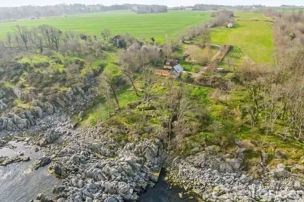 Hus ligger i en skovklædt kystområde omgivet af klipper; i baggrunden ses marker og skove.