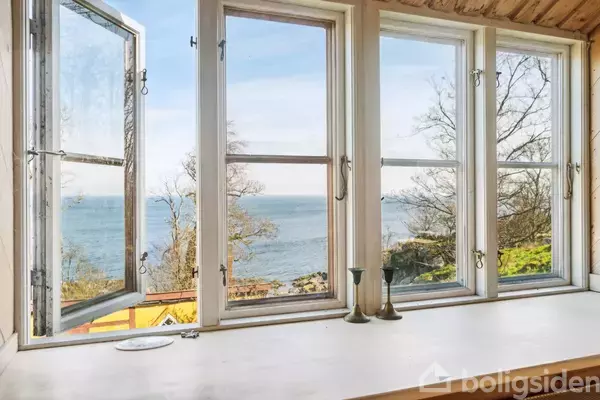 Et åbent, hvidt vindue med to lysestager står i en vindueskarm. Udsigt over havet og træer i baggrunden.