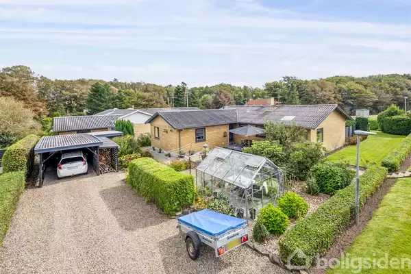 Et gult murstenshus med en grønalge drivhus i baghaven. Ved siden af huset er der en carport med en hvid bil, omgivet af velplejede hække og en trailer i forgrunden.