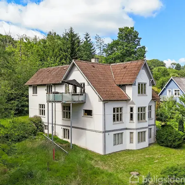 Hvidt hus med rødt tegltag står i en grøn have. Balkonen på siden er tilknyttet huset. Omkringliggende område er fyldt med træer og andre huse, med blå himmel i baggrunden.
