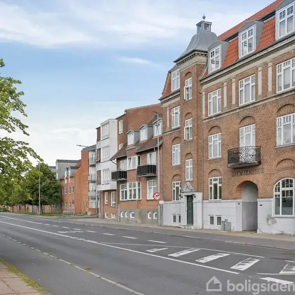 Rød murstensbygning står stille langs en tom gade med fortov. Træer står til venstre og skyet himmel ovenover i en bymæssig kontekst.