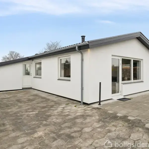 En hvid étplanshus med store vinduer står på en flisebelagt terrasse. Omkring huset er der en række træer og buske i baggrunden under en blå himmel.