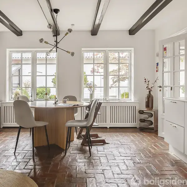 Rundt bord med fire stole omringet af et lyst rum. Brændeovn til venstre, store vinduer i baggrunden, og køkken til højre med grønne fliser. Lampe hænger over bordet.