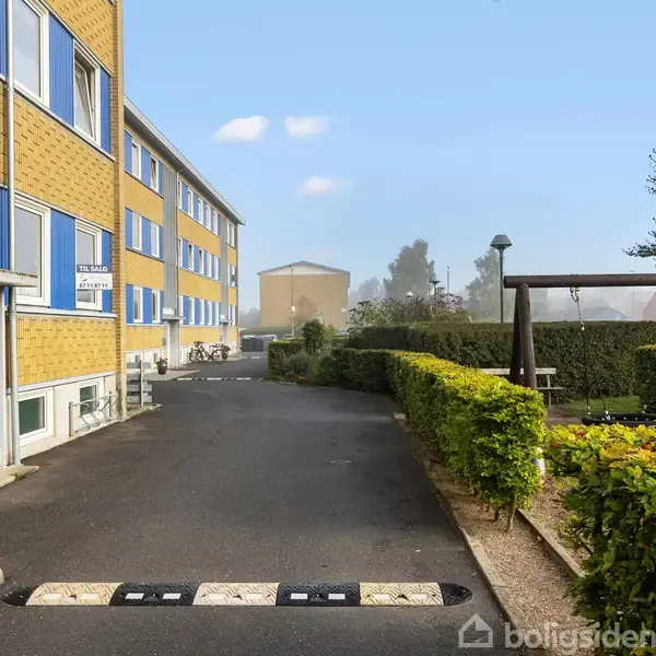 Et lejlighedskompleks med gule mursten og blå detaljer. En asfalteret sti fører forbi en gyngestativ mod en have. Der ses også skilte: "Praktisk Advokat Caitling Olsen" og "Cykelservice".