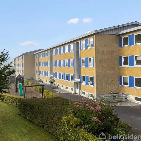 Et langt, gult murstenslejlighedskompleks strækker sig langs en vej, omkranset af en legeplads til venstre og en græsplæne med buske og træer i forgrunden.