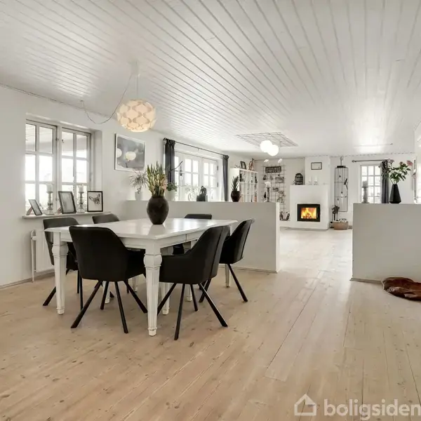 Spisebord med sorte stole står i en rummelig, lys stue med trægulv og hvidmalet loft. Hyldesystem og dekorationer præger væggene, mens en brændeovn yderst skaber hygge.