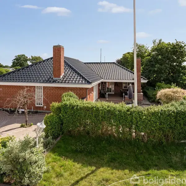 Rødt murstenshus med sort tag ligger i en velplejet have omgivet af buske og træer. Brolagt indkørsel strækker sig mod huset, og en flagstang står ved siden af.