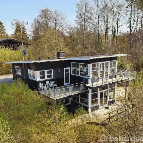 Et sortmalet træhus med stor terrasse står på en skovklædt bakke. Terrassen har en udsigt over omgivelserne, med en solrig himmel og grønne buske rundt omkring.
