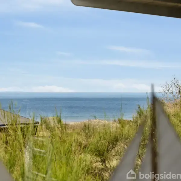 Kystlandskab set fra veranda, hvor små sejlformede dekorationer står i forgrunden. Havet og himlen mødes i horisonten, omkranset af grønne buske og et hus.