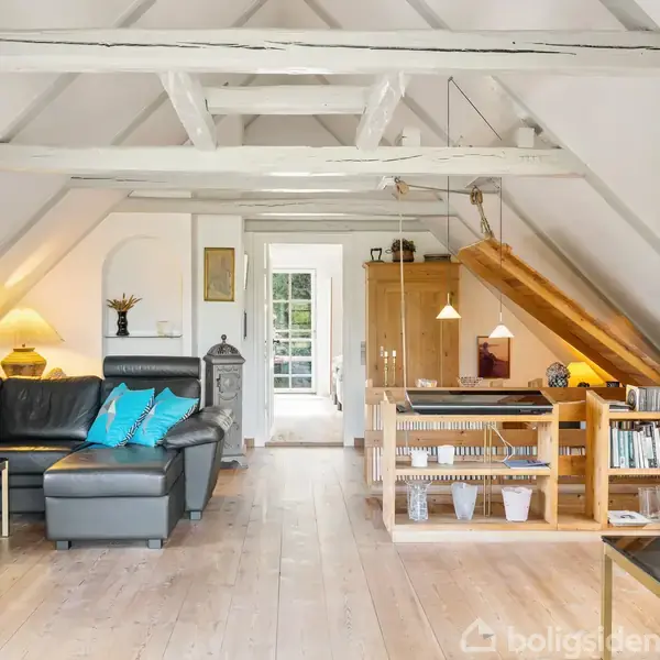 Stue med sort sofa, blå puder og sofabord står til venstre i et lyst rum med skrå lofter. Trætrapper fører op til hems over spisestue med trægulve.