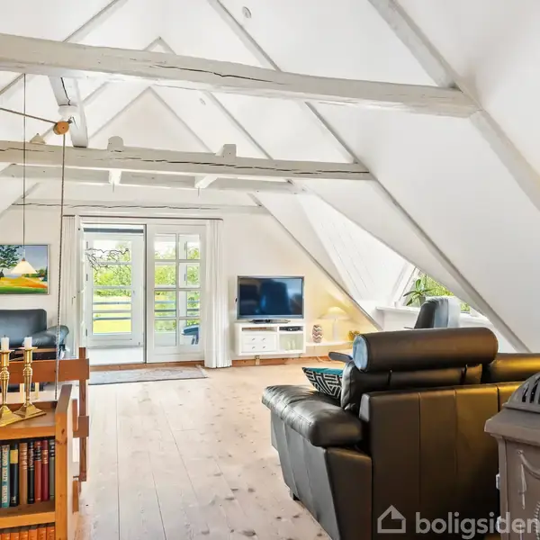 Stue med skrå vægge og hvidmalede loftsbjælker. Sorte lædermøbler står ved fjernsynet. Hylder med bøger og lysestager i forgrunden. Store vinduer giver udsigt til have.