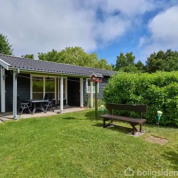 Et sort træhus med en overdækket terrasse og et bord med bænke, omgivet af en grøn have med buske, græsplæne og træer i baggrunden.