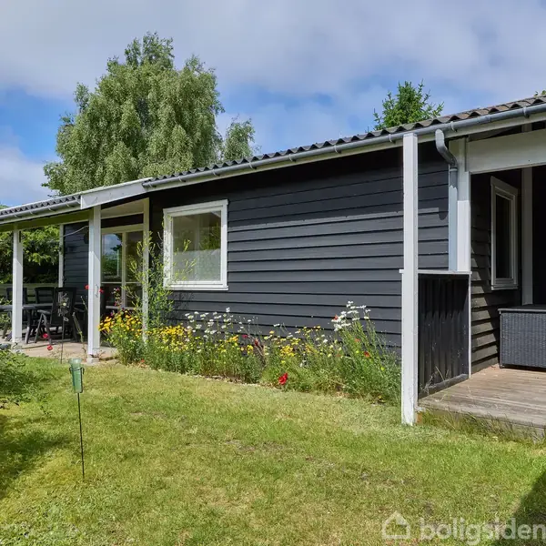 Et sort træhus med hvide vinduer står i en velplejet have. Blomster vokser langs facaden, mens en overdækket terrasse ses til venstre. Solrig himmel og grønne træer omkranser huset.