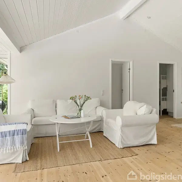 Stue med hvide møbler, herunder to sofaer og en lænestol med et rundt bord. Store vinduer til venstre slipper naturligt lys ind. Trægulv og lyse vægge skaber en lys atmosfære. En lampe hænger fra loftet i baggrunden.