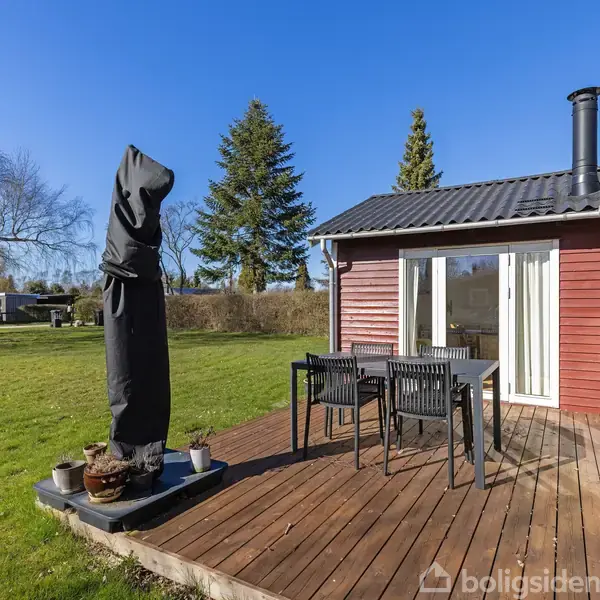 En terrasse med et bord og fire stole står foran et rødmalet hus med store vinduer. En overdækket grill eller parasol er placeret på terrassen. Græsplæne og træer i baggrunden.