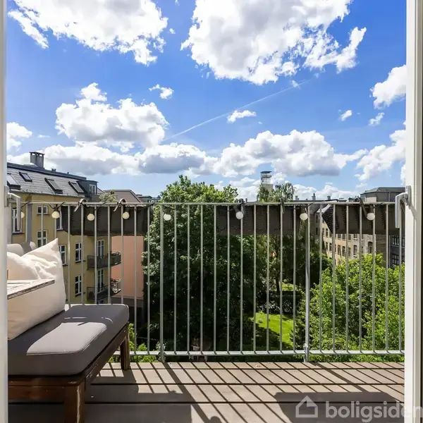 Åben altandør afslører en lille balkon med et sofabord og bænk. Udsigt over grønne træer og tagtoppe under en blå himmel med skyer.